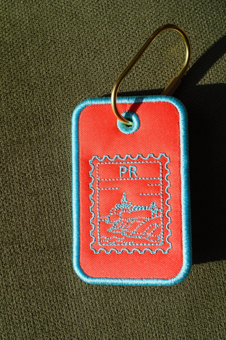 Orange Puerto Rico stamp embroidered luggage tag

Puerto Rican bag charm with gold-tone carabiner

Aqua embroidery detail on Puerto Rico travel tag

Puerto Rico luggage tag attached to a suitcase

Front and back of embroidered Puerto Rico bag tag