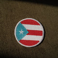 A circular embroidered patch featuring the Puerto Rican flag, placed on a textured olive green fabric background.