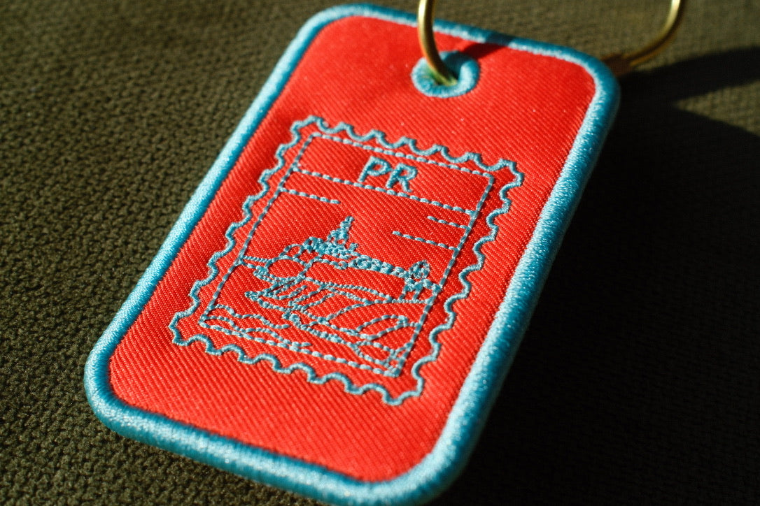 Orange Puerto Rico stamp embroidered luggage tag

Puerto Rican bag charm with gold-tone carabiner

Aqua embroidery detail on Puerto Rico travel tag

Puerto Rico luggage tag attached to a suitcase

Front and back of embroidered Puerto Rico bag tag